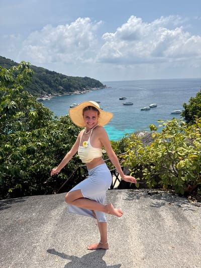 A young woman in Lam Kaen, Thailand stands on a stone surface with vegetation and the ocean as a backdrop. She wears a straw hat, a light-colored top with a flower accent, and a light-colored skirt or pants. She extends both arms outward. Boats are visible on the turquoise ocean. The sky is partly cloudy.