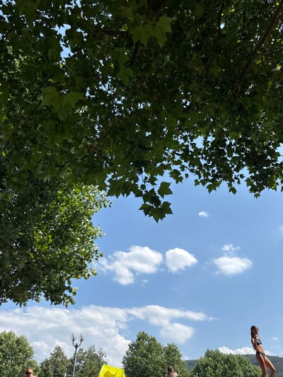 A sunny day with a bright blue sky and scattered white clouds is depicted. Lush green trees with large leaves dominate the upper portion of the frame, casting shadows. In the lower right quadrant, a young woman in a black bikini poses, looking slightly downwards. To her left, another person's back is visible, also seemingly dressed for warm weather. In the background, dense green trees line a hillside, and a modern street lamp with two globe-shaped lights stands centrally. A bright yellow object, possibly a tent or umbrella, is partially visible on the left side of the frame. The scene appears to be outdoors, suggesting a park or recreational area within Bürgstadt, Germany, likely during the summer.