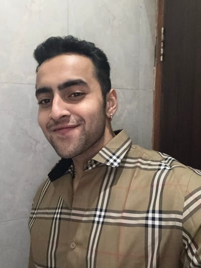 A man poses indoors, wearing a Burberry-patterned shirt. The shirt has a beige base with black, white, and red lines forming a plaid pattern. He has dark hair, an earring in his ear, and a slight stubble. The background includes a gray tiled floor and a dark brown wooden surface near the top. The image is rotated sideways.