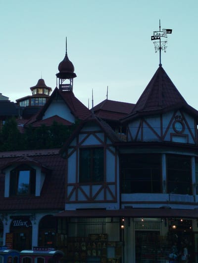 The image captures a twilight scene in Truskavets, Ukraine, showcasing a charming European-style building. The architecture features multiple red-tiled roofs, turrets, and decorative gables. A prominent weather vane, inscribed with "Club.PL.com" and "WWW", tops one of the spires, indicating a business or establishment. The building appears to be a commercial establishment, with a storefront visible at the base. Signage in Cyrillic script, reading "Шик", is displayed above an arched entrance, suggesting the name of the shop or business. The windows of the building are dark, reflecting the fading light. Displayed outside the storefront are various items, possibly souvenirs or crafts, arranged on shelves and tables, hinting at a market or retail area. The overall mood is calm and serene, fitting for the end of the day. The sky is a pale blue, typical of dusk. There are no clear indications of specific events, but the scene suggests a normal day of operation for the businesses.