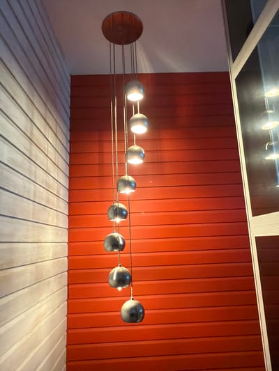 An indoor scene in Felixstowe, United Kingdom, depicts a modern multi-light pendant fixture suspended from a white ceiling. The fixture consists of a circular, metallic ceiling rose from which eight individual spherical, dark grey or metallic lamp shades hang at varying lengths via thin cables, forming a vertical cascade. Each shade emits an illuminated warm light source from its lower aperture. The background features two distinct wall surfaces meeting at a corner: a left wall clad in horizontal light-colored wooden slats, and a right wall clad in horizontal vibrant orange or red slats. To the far right of the frame, a vertical, light-framed reflective surface, likely a mirror or window, captures a partial, distorted reflection of the light fixture and the adjacent red wall. No individuals, specific actions, or interactions are depicted.