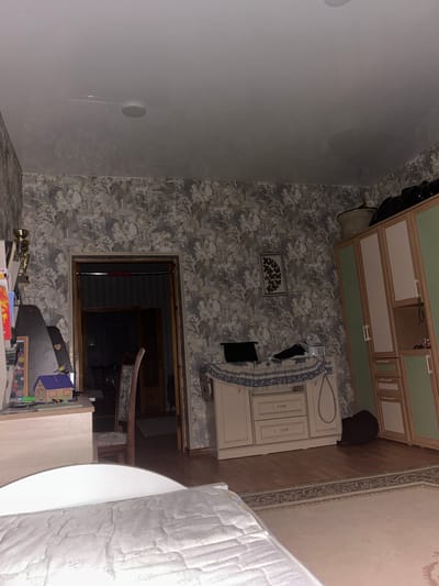 The image depicts a dimly lit bedroom with patterned wallpaper and wooden flooring. In the foreground, the edge of a white bed with a textured mattress is visible. To the left, a portion of a wooden dresser or desk with drawers is visible, topped with a small toy house and other assorted items. A tall, ornate chair with a patterned seat stands near an open doorway leading to another dark room. In the background, to the right, a tall wardrobe with light-colored doors and green accents is partially visible. A woven basket sits atop the wardrobe, and a black bag rests on the floor beside it. The overall impression is of a domestic interior, likely a bedroom, in Komsomolskoye, Russia, given the context provided. The lighting is low, suggesting it may be nighttime or the curtains are drawn.
