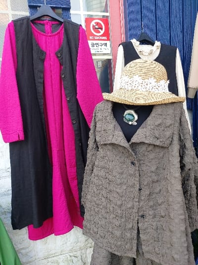 This image captures an outdoor clothing display, likely a storefront or market stall, in 강화군, South Korea.
On the left, a long black sleeveless vest with multiple black buttons hangs over a vibrant magenta long-sleeved dress or tunic. Both garments are suspended from a black hanger against a light background, partially obscured by a textured wall.
To the right, a mannequin torso displays a textured, crinkled grey long-sleeved jacket with a wide collar and three visible black buttons down the front. Resting on the jacket, a woven straw hat with a wide brim and white lace detailing sits atop a black sleeveless top, which is layered over a cream-colored long-sleeved shirt. A large green pendant necklace with a black cord is visible on the mannequin's chest, below the hat.
In the background, positioned between the two clothing displays, a red circular "NO SMOKING" sign with Korean text "금연구역" (Geumyeon-guyuk) is affixed to a building, confirming the location as South Korea. Above and to the right of this sign, blue vertical wooden panels are visible, possibly a door or shutter. A white window frame is also present behind the left display. The lower section of the image reveals a rough, light-colored stone or stucco wall texture. A small green fabric item is partially visible in the bottom left corner.