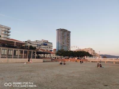 This image depicts a beachfront scene in Follonica, Italy, at dusk. The foreground is a sandy beach populated by several individuals and a baby stroller. To the left, a woman in a black dress and a child in a pink outfit are on the sand. Further back, a man sits on the sand next to a baby stroller, and another couple is seated on the sand, with one person holding a phone. In the middle ground, a raised promenade is lined with trees and buildings. One prominent building is a tall, blue high-rise with numerous balconies, and a Ferris wheel is visible behind it. To the left of the high-rise, there are other multi-story buildings. A restaurant with outdoor seating is visible on the far left, with people seated at tables. The background shows a white railing along the promenade and hills in the distance, under a pale blue sky with a hint of sunset glow. The "HUAWEI P30 Pro LEICA QUAD CAMERA" watermark is present in the bottom left corner.