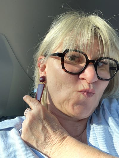 A female individual is depicted in a close-up, waist-up portrait taken inside a vehicle in the city of Orgon, France. The subject, who has fair skin and light blonde hair, wears dark-framed eyeglasses, revealing a light-colored left eye. Her mouth is pursed. On her left ear, she wears a dark red earring consisting of a spherical top piece connected to an elongated, rectangular lower section. Her left hand's index finger, adorned with a silver-toned ring, is positioned near the lower part of this earring. She is dressed in a light blue and white striped collared shirt. Strong sunlight illuminates the right side of her face and shoulder, creating prominent highlights and shadows. The light-colored interior roof lining of the vehicle is visible in the background.