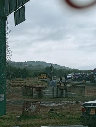 A statue stands on a raised platform, enclosed by a black fence, with people nearby. Vehicles line a road in the background, including a yellow bus, vans, and cars. A hill rises gently in the distance under a cloudy sky. A green pole with barbed wire and metal signs is in the foreground on the left. A weathered, cylindrical object is near the road's edge. Part of a dark sedan is visible at the bottom right.