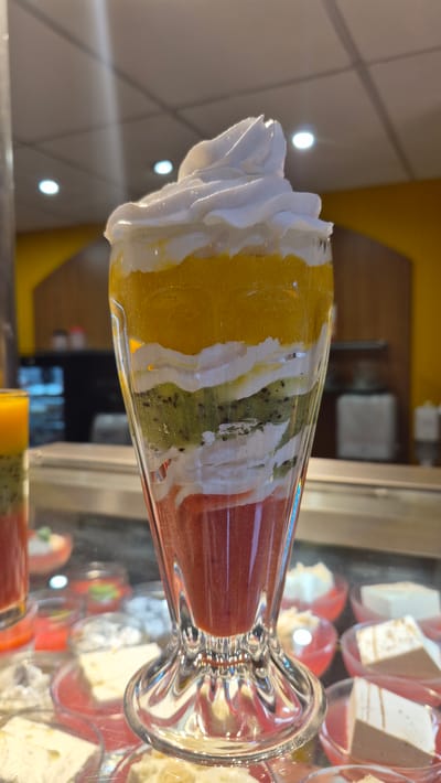 A tall, fluted glass dessert is filled with layers of fruit puree and whipped cream. The bottom layer appears to be a red fruit puree, possibly strawberry or raspberry. Above this is a layer of white whipped cream, followed by a layer of sliced kiwi. Another layer of whipped cream sits atop the kiwi, and then a thick layer of orange-colored fruit puree, likely mango or peach. The dessert is topped with a generous swirl of white whipped cream. The glass is clear and shows the distinct layers. The dessert is presented in what appears to be a display case, with other desserts and food items visible in the blurred background. The setting seems to be a food establishment, potentially a cafe or a dessert shop, with warm lighting and a yellow-colored wall in the background. The image does not contain any recognizable landmarks or clues that would indicate the location is specifically in Jēkabpils, Latvia.