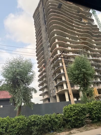 This image captures a multi-story concrete building under construction in Falomo, Nigeria. The structure features numerous floors with distinctive, undulating curved balconies. Scaffolding is visible across multiple sections of the building, and a dark construction safety net partially covers its left vertical expanse. A green construction crane boom is partially visible extending from the upper right of the building.

In the midground, a dark grey wall runs horizontally, delineating the perimeter of the construction site. A prominent brown utility pole, supporting multiple overhead wires, stands in the center-right. Green trees are positioned on the left and to the right of the utility pole, providing foliage cover. A dense green hedge occupies the lower portion of the frame. To the far left, a portion of a building with a reddish-brown tiled roof is visible behind the foliage. The sky above is partly cloudy. No individuals, direct actions, or interactions are visible within the static scene.