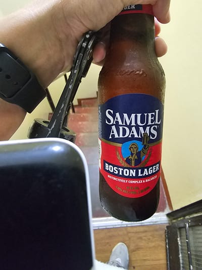 A hand holds a bottle of Samuel Adams Boston Lager. The bottle is brown with condensation. A multi-tool is being used to open the bottle. A black smartwatch is visible on the wrist of the hand holding the bottle. A black object, possibly a phone, is in the foreground and partially obscures the view. A wood floor is visible in the background. The location is Magdalena del Mar, Peru.