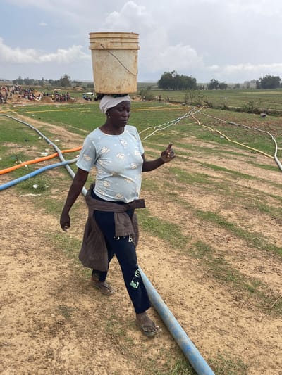 In an outdoor scene in Durumi, Nigeria, an individual stands on a dirt and grassy area. They balance a tan bucket on their head, and are dressed in a light blue t-shirt with a cartoon pattern, dark pants, and sandals. A white head covering is visible. The person is pointing. In the background, numerous people are clustered, and several hoses lay across the ground. The sky is overcast.