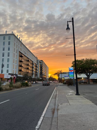The image captures an urban streetscape in Gainesville, Florida, USA, during either sunset or sunrise, with a prominent orange and yellow sky beneath scattered clouds. The view is along a paved, multi-lane road, likely looking west, given the direction of the intense light.

On the left side of the road, a modern, multi-story residential and commercial building, identified as "Midtown Village," features a white facade with numerous dark-framed windows and balconies. These surfaces reflect the vibrant sky. Ground-level commercial spaces, including a "Restaurant," are visible, fronted by a landscaped median and a row of parked cars.

The roadway features a light-colored pickup truck in the foreground, traveling away from the viewer, with other vehicles visible further down the street. White solid and dashed lines delineate the road and its shoulder.

On the right side, a concrete sidewalk runs parallel to the road. A tall, black, traditional street lamp post stands in the immediate foreground. Further along, a Chevron gas station sign is clearly visible, displaying fuel prices: Regular at $3.49, Plus at $3.99, and Premium at $4.39. Behind the Chevron sign is a building with "Bank of America" signage, and a large, green-leafed tree stands nearby. Other low-rise commercial structures are discernible in the distance on this side of the street. No people are visible. The overall scene depicts an active urban environment bathed in the warm light of the low sun.
