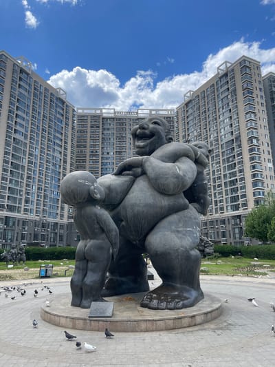 The image shows a bronze sculpture located in a park in Changping District, China. The sculpture depicts a man carrying other figures in a stylized, surreal manner. The man has a large grin and exaggerated features. The figures he carries are intertwined and indistinct. The sculpture sits on a circular platform surrounded by gray pavers. Several pigeons are on the ground near the base of the sculpture. Behind the sculpture are tall apartment buildings with a clear blue sky and some white clouds. Some green foliage is visible to the right of the buildings.