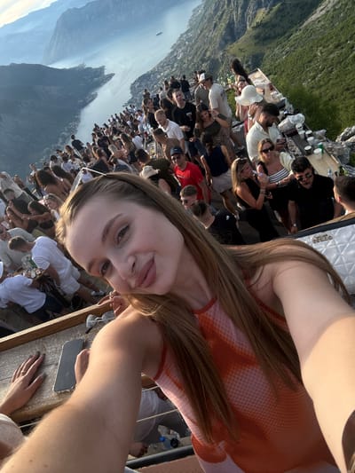 A group of people are gathered on a raised wooden platform overlooking a bay in a mountainous region near Žanjev Do, Montenegro. The platform, equipped with tables and chairs, appears to be part of an outdoor dining or event space. In the foreground, a woman is taking a selfie, with a blurry view of her arm and shoulder. Behind her, a large crowd of men and women are socializing, some standing, others seated at tables laden with drinks and food items. The background features a body of water, possibly a bay or fjord, with steep, green mountains rising on either side. The sky is clear with some hazy atmospheric conditions, suggesting it is daytime, possibly late afternoon due to the golden light. The overall atmosphere seems festive and relaxed.
