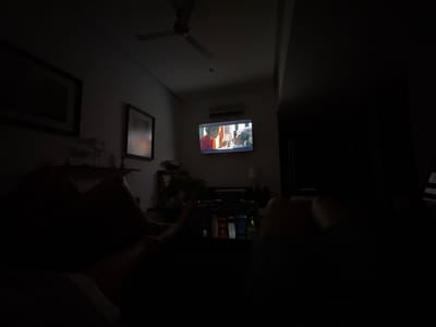 The image shows a dimly lit interior space, possibly a living room, with a television as the focal point displaying a program with two figures. The low lighting obscures most details, but one can discern what appears to be an air conditioning unit mounted above the television. The ceiling fan is visible, and there are frames hanging on the wall. In the foreground, there are vague outlines of what appear to be people sitting, possibly on a couch. A table with objects is located in front of the people.
It's not possible to pinpoint the exact location in Puerto Marqués, Mexico based on this picture.