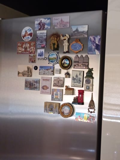 A close-up, eye-level shot shows a collection of novelty magnets adhered to a stainless steel refrigerator door. The magnets are varied in shape and depict miniature scenes, landmarks, and humorous figures, many appearing to be souvenirs from various global locations. Several magnets showcase architectural details and cityscapes, with some suggesting European locations such as Baden, Austria, indicated by a magnet featuring the "Baden" text and a recognizable building. Other magnets include a troll figure, a person in a chef's hat, a red British-style telephone box, and various miniature houses and buildings. The overall impression is of a well-traveled individual with a penchant for collecting mementos from their journeys.