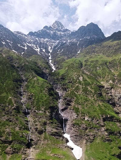 The image displays a breathtaking view of a mountain range, possibly in the vicinity of Ropsang, India. The mountains are a dramatic spectacle of towering peaks, some capped with patches of snow, even amidst the lush green slopes that dominate the lower and mid-altitudes. Several waterfalls cascade down the steep inclines, their white streams contrasting sharply with the dark rock faces and vibrant vegetation. The sky above is a canvas of white clouds scattered across a pale blue expanse, suggesting a clear, possibly cool, day. The rugged terrain and the presence of snow indicate a high-altitude environment, characteristic of the Himalayan regions where Ropsang is located. There are no people or man-made structures visible in the frame, emphasizing the wild and untouched nature of the landscape. The scene is one of raw, natural beauty, capturing the grandeur of the mountains, the dynamic flow of water, and the resilience of vegetation in a challenging environment.
