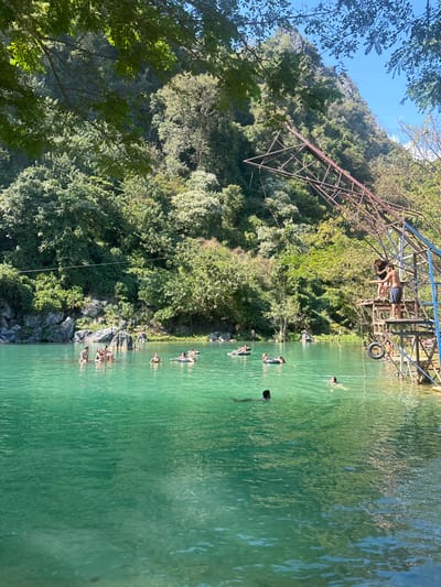 A turquoise lake is bordered by lush green vegetation and a mountainous backdrop. A metal structure with a ladder stands above the water, where a person is poised to jump. Several people are swimming and floating in the water. Sunlight reflects on the lake's surface, creating a shimmering effect.