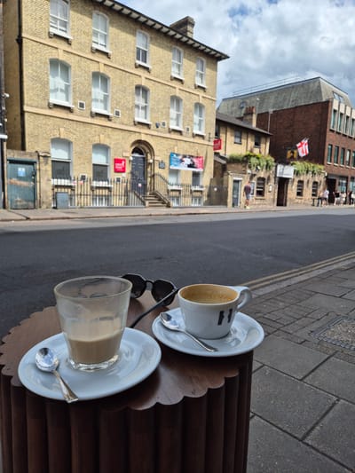 The image shows a view from a table outside a cafe in Cambridge, UK, with two drinks and a pair of sunglasses on the table. In the foreground, there is a wooden table with a scalloped edge. On the table are two white saucers, each with a spoon. One saucer holds a nearly empty glass of a milky coffee drink, and the other holds a white coffee cup with a brown, foamy coffee, a logo with the letters "H" is visible on the cup. A pair of dark sunglasses rests between the two drinks.
In the background, across a street, are buildings. To the left is a pale yellow brick building with multiple windows. To the right is a darker brick building with a sloped roof and windows. A red and white flag, possibly the St. George's Cross, is visible on the darker building. Several people are walking on the sidewalk in front of the buildings. The scene appears to be a street-level view from a cafe situated on a sidewalk in Cambridge, UK.