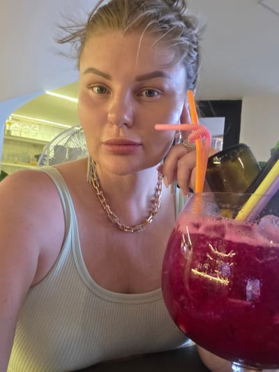 A close-up, waist-up, selfie-style portrait shows a woman with blonde hair pulled back and a light-colored tank top. She is wearing a thick gold chain necklace and has a large, red-colored beverage in a round glass with straws and a dark bottle in the background. Her left hand is resting on her chin, and she is wearing rings. The background appears to be indoors with shelves and a fan visible. The image does not provide any context or information about the location in Jiyang District, China.