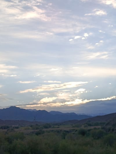 The image depicts a vast, cloudy sky over a mountainous landscape. The sky is filled with soft, wispy clouds in various shades of white, light blue, and pale pink, suggesting either sunrise or sunset. Sunlight filters through some of the clouds, creating streaks of light that descend towards the horizon.
Beneath the sky lies a series of rolling hills and mountains, appearing in darker, muted blue and purple tones. The closest hills are covered in scrubby vegetation and sparse green foliage. A few faint, darker vertical lines, possibly utility poles, are visible in the mid-ground. The furthest mountains are silhouetted against the sky, with subtle variations in their contours. The overall impression is one of a remote and expansive natural environment, likely during transitional light conditions of dawn or dusk. The scene is devoid of any human figures or explicit man-made structures, emphasizing the natural elements of sky and terrain. The location in Naryn, Kyrgyzstan, suggests a high-altitude, possibly steppe or mountainous region.