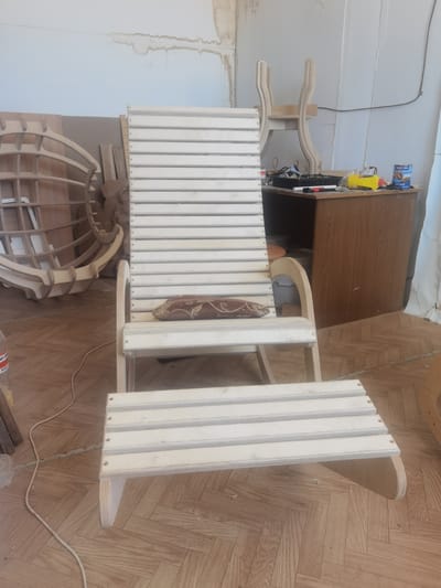 This image shows a wooden chaise lounge and footrest, seemingly unfinished, in what appears to be a workshop or garage. The chaise lounge is made of light-colored wood, with a slatted back and seat. A small, patterned cushion rests on the seat. The footrest is also made of slatted wood and is positioned in front of the chaise.
In the background, there are other wooden furniture pieces in various stages of completion. To the left, a curved, lattice-like wooden structure is visible. To the right, a wooden desk or cabinet stands against a wall. On top of the desk are various tools and supplies, including a can of what appears to be wood stain or varnish with Russian text that includes "ПРЕСТИЖ" and "ЯХТНЫЙ". A yellow spray can and some tubes of material are also present. The walls appear to be concrete or plaster, with some water stains visible on the upper left wall. The floor is made of parquet wood.
The overall impression is one of a work in progress, with a focus on woodworking. There are no people visible in the image. The lighting suggests it could be daytime, but the indoor setting makes it difficult to determine the time of day or weather conditions. The location is identified as Cherkessk, Russia.