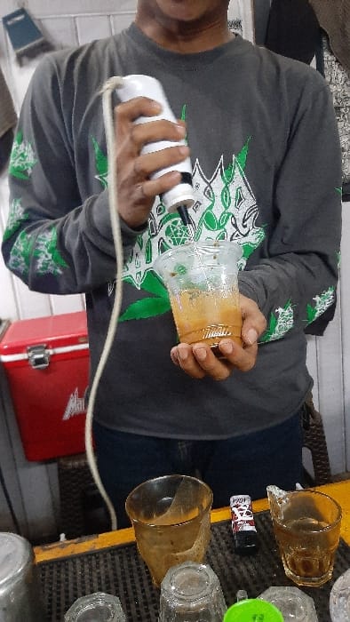 In RawangItiek, Indonesia, an individual is depicted indoors, likely at a beverage stall. The person, whose torso and lower face are visible, wears a dark grey long-sleeved t-shirt with green and white cannabis leaf patterns on the sleeves and a complex design on the chest, paired with dark blue jeans. They are actively mixing a light brown, frothy beverage in a clear plastic cup with a white and black battery-operated hand-held frother, holding the cup with their left hand and operating the frother with their right. A light-colored cord extends from the frother. The immediate foreground features a black ribbed mat on a yellow surface, displaying various items including a silver metal container, a tall amber glass, a black lighter with red and white branding, a smaller amber glass, and several clear, empty glasses. In the background to the left, a red cooler with a silver latch is visible. A dark brown woven stool is partially visible to the right. A blue and grey cleaning tool hangs on a white wall surface in the upper left.