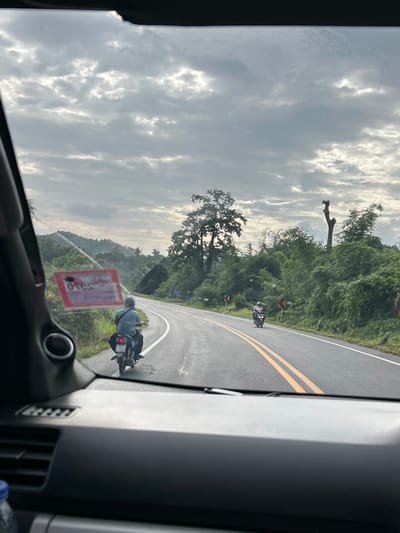 This is an over-the-shoulder shot from inside a vehicle looking out the windshield. The scene depicts a two-lane asphalt road curving to the left, bordered by lush green vegetation and rolling hills. The sky is overcast with dramatic cloud formations.
Two motorcyclists are visible on the road. The first, closer to the camera and in the left lane, is a person wearing a striped shirt and dark pants, riding a motorcycle with a license plate. They are facing away from the camera and heading down the road. On the right side of the road, further in the distance, a second motorcyclist is seen, dressed in pink and riding a motorcycle.
On the left side of the road, a red sticker with text and graphics is affixed to the windshield of the vehicle the photo is taken from. The surrounding environment suggests a rural or semi-rural setting, possibly in or near Ban Bang Hin, Thailand, given the vegetation and road infrastructure. The overall impression is a travel scene on a cloudy day.
