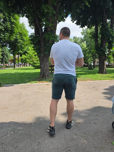 An adult male is observed from the rear, standing on a paved path within a park in Elin Pelin, Bulgaria. He is dressed in a white short-sleeved t-shirt, dark green athletic shorts, and black and olive green sneakers. His arms are positioned behind his back. The grey path is scattered with leaf litter and small organic debris. A partial view of a light blue item and a wheel, likely from a stroller, is visible to the man's immediate right. The scene's midground features a manicured green lawn and numerous large trees with dense foliage, indicating a summer environment. Sunlight projects shadows onto the path. In the background, additional trees and architectural structures are visible, consistent with a public park situated within a town or city. Distant figures are faintly discernible on the far left. The sky, seen through the tree canopy, suggests a bright day with minimal cloud cover.