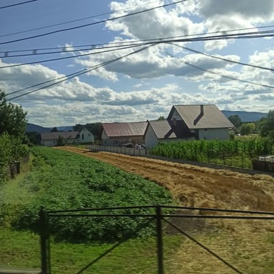 The image depicts a rural landscape in Vicovu de Sus, Romania, under a sky filled with white, fluffy clouds against a bright blue backdrop. Several power lines stretch across the top of the frame, casting shadows. In the mid-ground, a collection of houses with tiled roofs are visible, some featuring white or light-colored walls. One house has a prominent chimney. Adjacent to the houses, there are fields. One field is covered in lush green vegetation, possibly crops, while another appears to be harvested or is a field of dry straw. A dark metal fence runs horizontally across the foreground, partially obscuring the view of the fields and casting shadows. Trees and hills are visible in the distance. The overall impression is of a serene, sunlit day in a Romanian village.