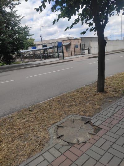 The image captures a street scene in Kostopil, Ukraine. In the foreground, a sidewalk paved with interlocking bricks is visible, with a damaged manhole cover set into the ground. Adjacent to the sidewalk is a strip of dry, yellowed grass. A tree with a dark, textured trunk stands on the right side of the image, its leaves casting shadows. The road, marked with white lines, runs horizontally across the middle of the frame. On the left side of the road, a metal fence encloses an area where numerous bicycles are parked under a covered structure. Behind the fence, a small, brick building with a metal door and a sign is situated. Further in the background, there are larger industrial-looking buildings and a long, white wall. The sky above is partly cloudy.