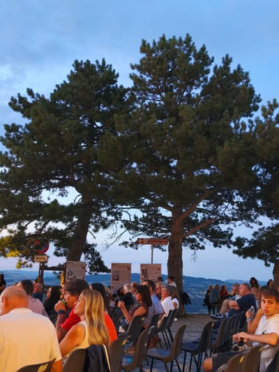 This is a medium shot photo of a crowd of people sitting in chairs outdoors, facing away from the camera, and towards a distant landscape under a twilight sky. The people are of various ages and genders, dressed in casual clothing. Many are looking towards something out of frame, suggesting an event or performance. Two large pine trees dominate the foreground, their branches casting shadows over the scene. There are signs in Italian indicating directions to "Ruderl del Castello Normanno-Svevo" and a parking restriction. The backdrop is a rolling, hilly landscape with distant mountains. The overall atmosphere is one of a relaxed outdoor gathering. The sign pointing to "Ruderl del Castello Normanno-Svevo" indicates that this scene is likely located in Bojano, Italy, as this refers to the Norman-Swabian Castle ruins in that town.