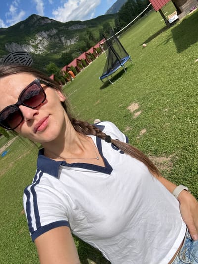 A woman with long brown hair in a braid, wearing a white polo shirt with blue trim and sunglasses, takes a selfie outdoors. She is in front of a grassy area with a volleyball net, a trampoline, and a row of red-roofed buildings. In the background, a mountain range is visible under a blue sky with clouds. The scene appears to be a resort or recreational area in Gusinje, Montenegro.