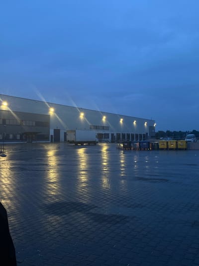 The image depicts a large industrial building with multiple loading docks and lights illuminating the wet, paved area in front of it. Several rows of large colored bins (blue, yellow, and green) are lined up near the loading docks. A white truck is parked at one of the loading docks, with its back doors open. The sky is a deep blue, suggesting either early morning or late evening, and the lights from the building reflect on the wet ground, creating a shimmering effect. The location is identified as Bielany Wrocławskie, Poland. There are no discernible people in the image. The scene is static, with no apparent actions or interactions taking place.