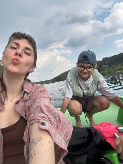 In the foreground, a woman with dark hair is captured in a selfie, puckering her lips. She is wearing a pink and white plaid shirt over a brown tank top, with a silver necklace featuring a small cross. Her left arm, visible in the lower left corner, has a tattoo and several moles. Beside her, a younger person wearing glasses, a dark blue baseball cap, and a green life vest is crouching. This individual is wearing a grey t-shirt with text that reads "LOVE ME AS I AM" and dark shorts. They are both in a bright green vessel on a body of water. In the background, a wooded shoreline with scattered trees and some distant figures can be seen under a cloudy sky. The image appears to be taken on a lake or river, possibly in the city of Dzerzhinsky, Russia, given the mention of a specific location.