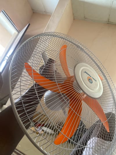 The image shows a close-up, low-angle view of an electric fan. The fan's blades are orange, and the protective grille is made of silver-colored metal wire. The central hub of the fan is white and features a circular logo with the text "EASY POWER" written around it in blue, along with a stylized "O" inside a circle. The fan appears to be indoors, and the background shows a beige-colored wall and ceiling, with some ceiling fixtures visible. There is also a glimpse of a window or opening in the lower left of the frame. The lighting suggests it is daytime. The overall scene is mundane and domestic, with no specific activity clearly depicted beyond the presence of the fan. The focus is primarily on the fan itself.