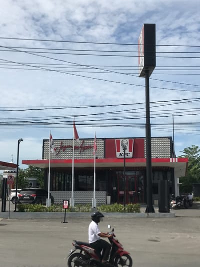 The image depicts an outdoor scene in Lhoksukon, Indonesia, featuring a KFC fast-food restaurant under a partly cloudy sky during daylight hours. The KFC building is characterized by its red and white facade, accented with a grid-patterned upper section. A tall signpost with the iconic red and white KFC logo, including Colonel Sanders and the "KFC" text, stands prominently near the building.
Numerous black power lines stretch across the upper portion of the frame. In the foreground on the right, a person wearing a white shirt and a black helmet is seated on a red and black scooter or motorcycle, facing away from the viewer. The ground is paved, resembling a road or parking lot.
Several small red and white flags are positioned in front of the restaurant. One flag clearly displays Arabic script which reads "لا إله إلا الله" (La ilaha illallah - There is no god but Allah). Another flag appears to have Latin script that is not entirely legible. A "No Parking" sign, denoted by a circled 'P' with a diagonal line through it, is visible on the paved area. In the background, a black SUV and another motorcycle are parked or moving.