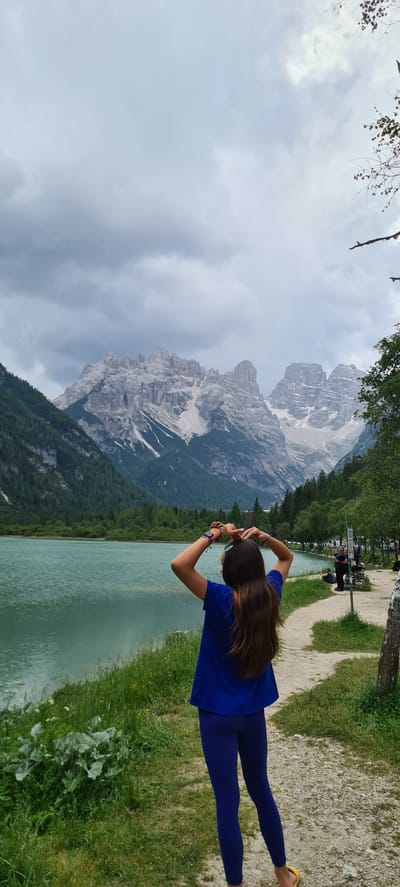 A vertical, medium shot depicts a young woman facing away from the camera, with her hands raised and clasped above her head, forming a heart shape. She is positioned on a dirt path next to a serene, turquoise lake. The woman has long, brown hair and is wearing a vibrant blue long-sleeved shirt and blue leggings. Her left wrist bears a watch and a scrunchie.

The background showcases a breathtaking mountain landscape. Jagged, rocky peaks with patches of snow dominate the horizon under a cloudy, grey sky. Lush green pine forests cover the lower slopes of the mountains. The lake's surface is calm, reflecting the overcast sky and surrounding greenery.

To the right of the path, a few individuals are visible in the distance, some sitting and others standing. A tree trunk is situated on the right edge of the frame. The overall scene captures a moment of quiet contemplation or appreciation of nature's grandeur in the vicinity of Toblach-Dobbiaco, Italy.
