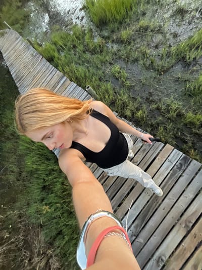 The image shows a fair-skinned female with shoulder-length blonde hair, wearing a black tank top and light-colored pants, standing on a wooden boardwalk. She is holding a small black electronic device in her left hand, presumably taking the selfie. The boardwalk appears to be adjacent to a grassy, marshy area with standing water. The location is Khwai, Botswana. The individual is wearing multiple bracelets on her left wrist.