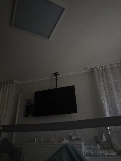 The image shows an interior view, likely a bedroom or hospital room. The scene is dimly lit, with a large, dark television screen mounted on the wall and a ceiling fixture visible at the top. A patterned curtain hangs to the right, and a framed picture of two people is on the wall to the left of the television. Below the television, a dresser or cabinet is visible with various items on top, including bottles and what appear to be tissues. A dark, horizontal bar is in the foreground, obscuring part of the lower section of the scene. The overall impression is one of a domestic or institutional setting. The location is specified as Заплавская, Russia.