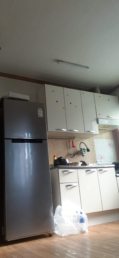 The image shows a kitchen scene. A silver refrigerator is on the left side of the frame. Above a kitchen counter, there are white cabinets adorned with small heart-shaped stickers in red, yellow, and pink. A silver range hood is installed below some of the cabinets. A faucet with a curved neck is mounted on a tiled backsplash. On the counter, there is a black pot, a yellow bottle, a spatula, and a bowl. To the right of the refrigerator, on the floor, is a clear plastic bag containing items, one of which appears to be a Pepsi bottle. The floor is made of wooden planks. The ceiling features a long fluorescent light fixture. The image appears to have been taken indoors, in a residential kitchen. The presence of a refrigerator, cabinets, sink, and stove area indicates it is a functional kitchen space. The heart stickers suggest a decorative touch, possibly for a holiday or personal preference. The lighting in the room seems to be a combination of overhead artificial light and natural light entering from an unseen source, casting shadows. The overall impression is of a tidy, albeit somewhat dated, kitchen.