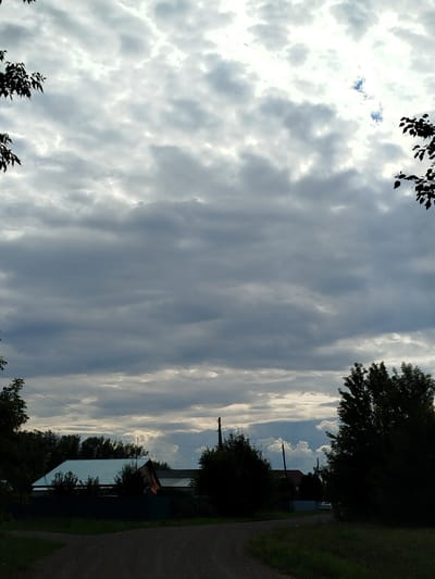 A low-angle outdoor shot captures a cloudy sky above a rural area. The sky is filled with numerous gray clouds, with some lighter patches indicating the sun's presence. In the lower portion of the image, a road leads towards a few residential buildings and trees. The buildings have dark-colored roofs and are surrounded by vegetation. A power line is visible stretching across the scene. The overall impression is of a natural, somewhat overcast day in a village setting.