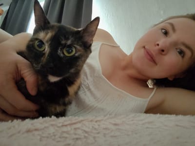 A young woman with light skin and brown hair pulled back is lying down, holding a tortoiseshell cat. The cat is looking directly at the camera with bright green eyes. The woman is wearing a white tank top with a delicate lace pattern. They are both lying on a soft, light pink, fuzzy surface, possibly a blanket or bedspread. The background shows a wall with textured, possibly painted, finish and a dark curtain.

The overall mood of the image is relaxed and intimate, suggesting a casual moment between the woman and her pet. The lighting appears to be natural, coming from a window, and the time of day is likely daytime. There is no visible text in the image. The setting appears to be indoors, possibly a bedroom or living room. Given the location context of Korosten, Ukraine, this could be a domestic scene within a home in that region.