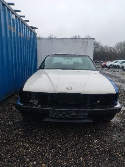 This image captures a front-facing view of a heavily damaged sedan, primarily white with blue front fender sections, positioned outdoors on a dark, wet gravel surface. The vehicle's front grille and headlights are absent, exposing the radiator and other internal engine bay components. The white hood exhibits stains and dirt. A black plastic front bumper and lower valance are present. The windshield is intact, reflecting the grey, overcast sky, and the vehicle's interior shows dark seats and a hanging air freshener. To the immediate left of the sedan, a weathered blue corrugated metal shipping container stands. In the background, beyond a light-colored concrete wall, bare trees are visible, and an auxiliary vehicle storage area contains multiple parked vehicles, including light-colored cars and vans, some appearing dirty. No individuals, actions, or interactions are depicted. The scene suggests an industrial yard or an auto salvage facility. The specific location within Bitburg, Germany, is not discernible from the image's content.