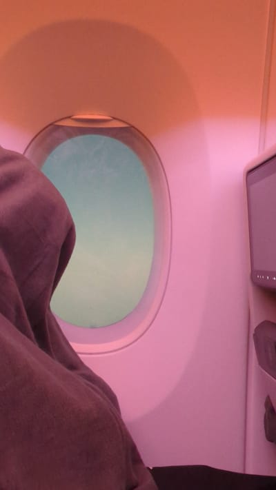 The image shows an interior view from an airplane seat. A dark purple blanket is draped over a person's lap, partially obscuring the window. The airplane window is oval-shaped and shows a hazy, light blue sky outside. The interior of the airplane cabin is illuminated with a pinkish-orange light, creating a warm ambiance. To the right of the window, a personal entertainment screen is visible, mounted on the back of the seat in front. The screen appears to be turned off, and the surrounding cabin wall is painted a light pink color. The scene is devoid of any specific city or location markers, and thus the city is unknown.