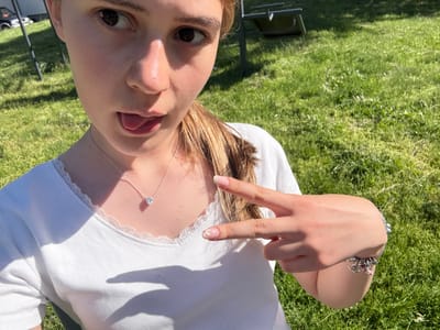 A fair-skinned person, likely female, is lying on her back in an outdoor grassy area, possibly in Panagyurski kolonii, Bulgaria, as per the prompt. She is wearing a white top with lace trim and a silver necklace with a heart-shaped pendant. She is holding up her left hand in a "peace" sign, and her nails are decorated with distinct designs. Her face is visible, with her tongue sticking out slightly. There is a bracelet with charms on her left wrist. The background consists of green grass and shadows cast by sunlight. There is also a chair visible in the background.