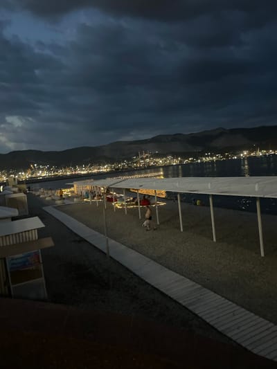 The image depicts a nighttime scene on a beach in Neberdzhaevskaya, Russia. The sky is dark and cloudy, with a hint of twilight. In the distance, a city skyline is illuminated by numerous lights, suggesting an active port or industrial area. The foreground features a beach with a wooden boardwalk leading towards a covered area. Several people are visible, some seated on cushions under the canopy, while others are walking around. The area is decorated with string lights, creating a festive atmosphere. The sea is dark and calm. The overall impression is one of a lively beachside gathering at dusk or early evening.