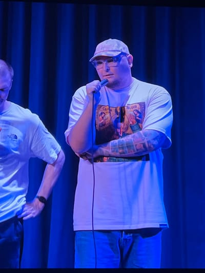 A man wearing a light-colored baseball cap, glasses, and a white t-shirt with a religious graphic and a tattooed arm is holding a microphone. He is standing in front of a dark blue curtain. Another man in a white t-shirt with "The North Face" logo is partially visible to the left, holding the first man's hand. The lighting is predominantly blue. The location is a stage in the city of Genemuiden, Netherlands.