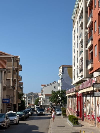 This is a street-level, vertical, full-body shot of a residential and commercial street in Lapčići, Montenegro. The street is lined with multi-story buildings, some with arched balconies, and features a clear blue sky. On the left, a building with terracotta-tiled roofs and beige facades houses shops, one labeled "ADMIRAL JET." On the right, a modern, tall building with white and orange facades is prominent, featuring shops at street level with large advertisements and a sign reading "AKTIVA." A few cars are parked along the street, including a white taxi. Several people are visible walking on the sidewalks, some browsing in front of the shops. A woman in a white top and pink shorts walks towards the viewer on the right sidewalk. The street is moderately busy with pedestrian and vehicle traffic.
