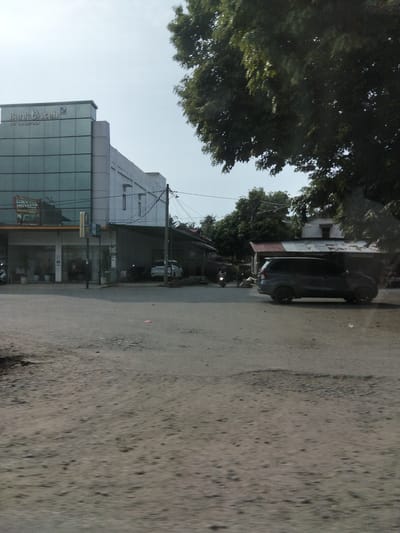 The image captures a street scene in Keudelapang, Indonesia. On the left, a modern, multi-story building with a glass facade dominates the frame. The building's upper portion displays the "Bank Aceh" logo. Below the bank, a commercial storefront is visible with potted plants and an awning. A traffic light stands in front of the building. To the right, a dark-colored SUV is parked on a dirt area next to a structure with a corrugated metal roof. In the background, trees and other buildings suggest a semi-urban environment. The road surface appears to be unpaved. A motorcycle is seen on the road in the middle distance, moving away from the camera. Power lines are visible stretching across the scene. The sky is overcast.