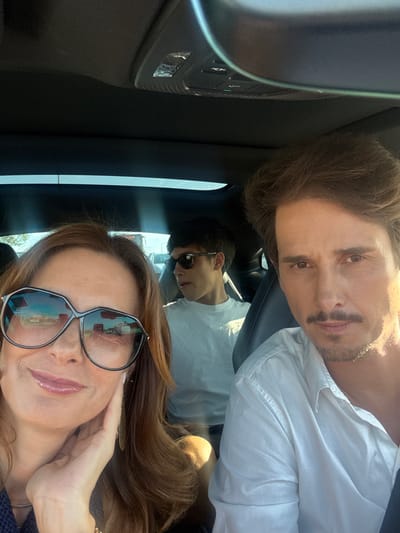 This media file is a selfie taken inside a car, showing three people. The person in the foreground is a woman with long brown hair, wearing large, dark-rimmed glasses and a navy blue top. She is touching her face with her right hand, and her lips are shiny and red. Behind her, a man with brown hair and a mustache is visible, wearing a white shirt. He is looking towards the camera. In the middle ground, a younger person with short dark hair is also looking towards the camera, wearing sunglasses and a white t-shirt. The setting appears to be a sunny day, as indicated by the bright light and reflections in the woman's glasses, which show a sky and possibly buildings. The interior of the car is visible, with dark upholstery and part of the car's ceiling console with lights. The orientation of the photo suggests it was taken vertically. The overall mood seems casual and happy. There is no visible text.